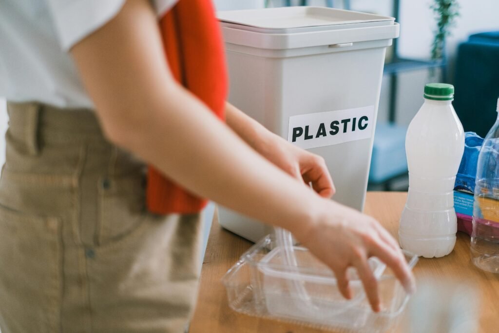 Crop anonymous female in casual clothes standing near bucket for plastic and sorting out rubbish in light room showing plastic waste management